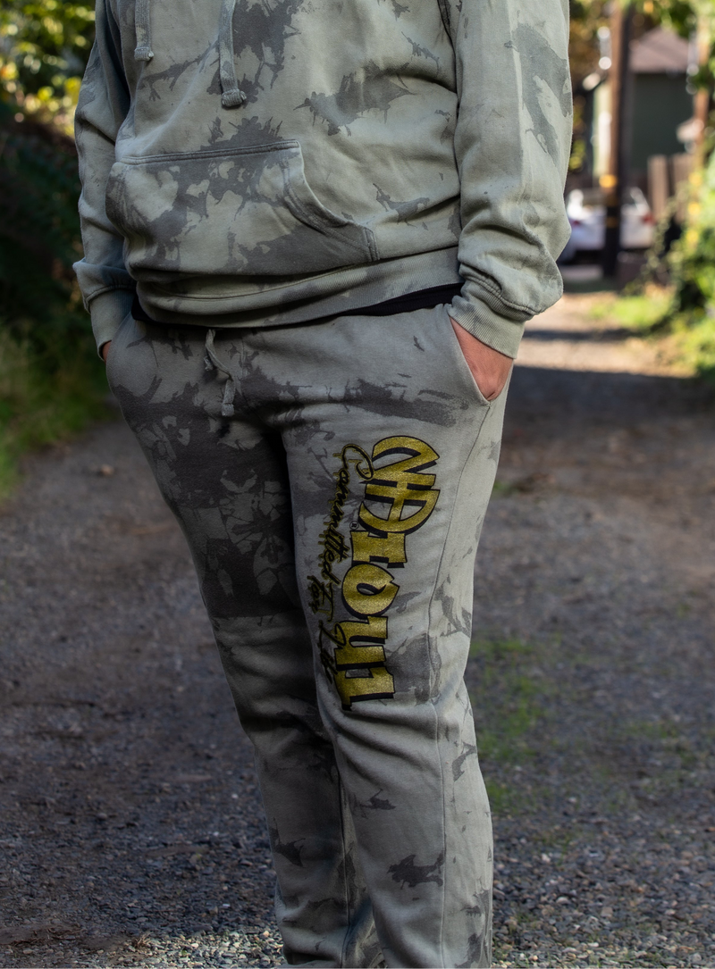 Load image into Gallery viewer, Person wearing camouflage hoodie and pants with a logo on a gravel path.