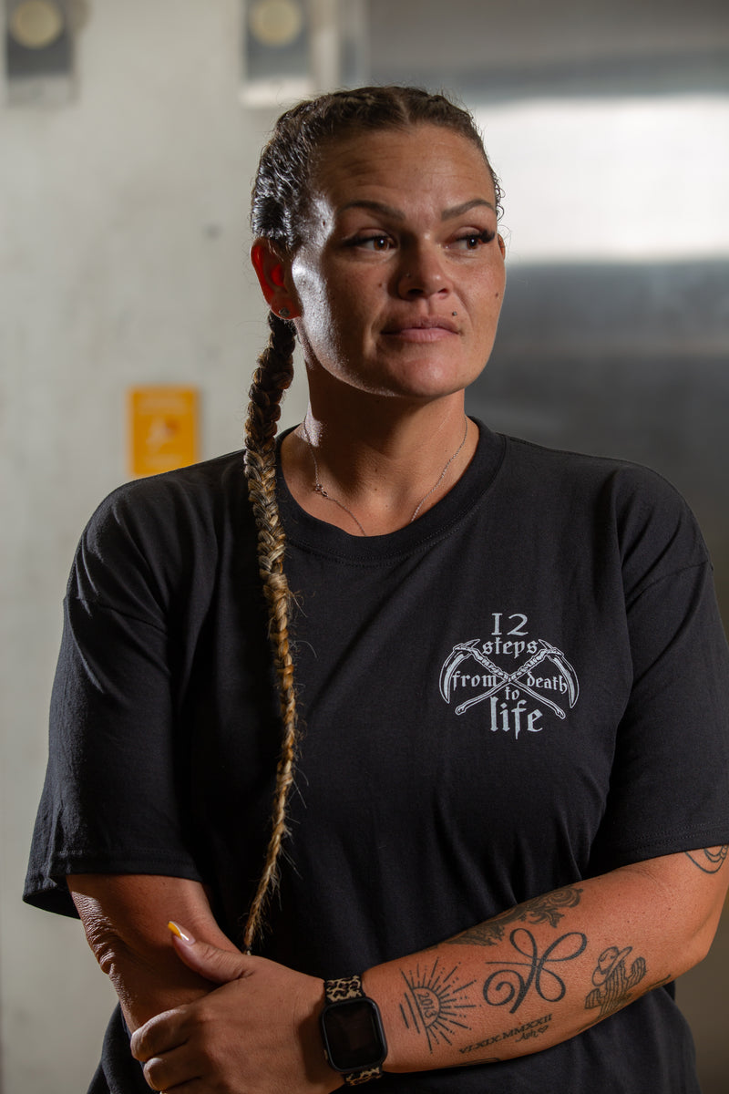 Load image into Gallery viewer, Person wearing a black t-shirt with a logo and text, standing against a plain background.