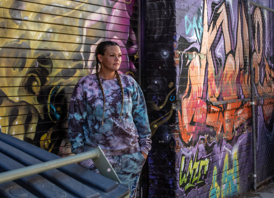 Person wearing a tie-dye hoodie and pants standing in front of a graffiti-covered wall.