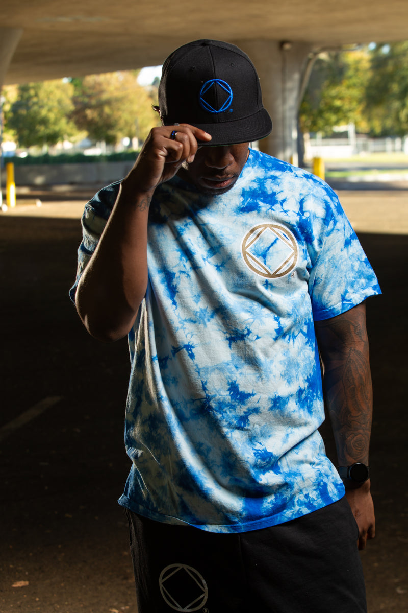 Load image into Gallery viewer, Person wearing a blue tie-dye shirt and black cap with a logo, standing outdoors.
