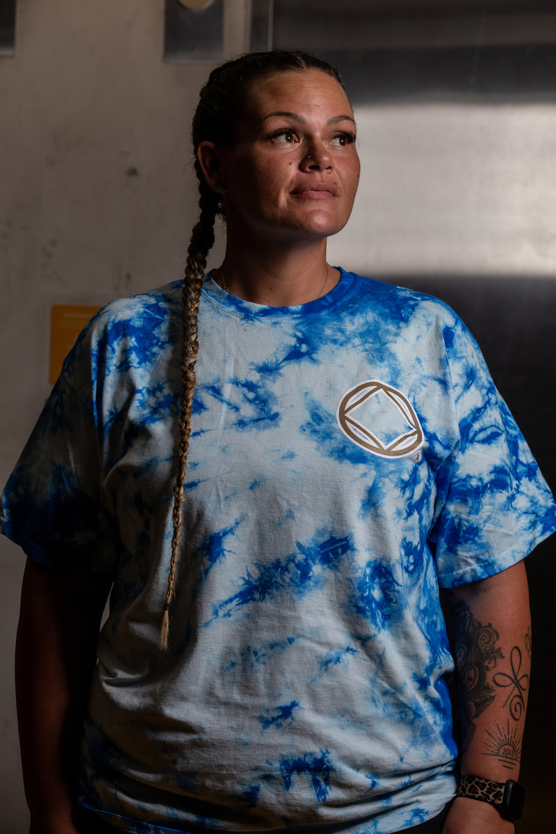 Load image into Gallery viewer, Person wearing a blue and white tie-dye t-shirt with a logo, standing indoors.
