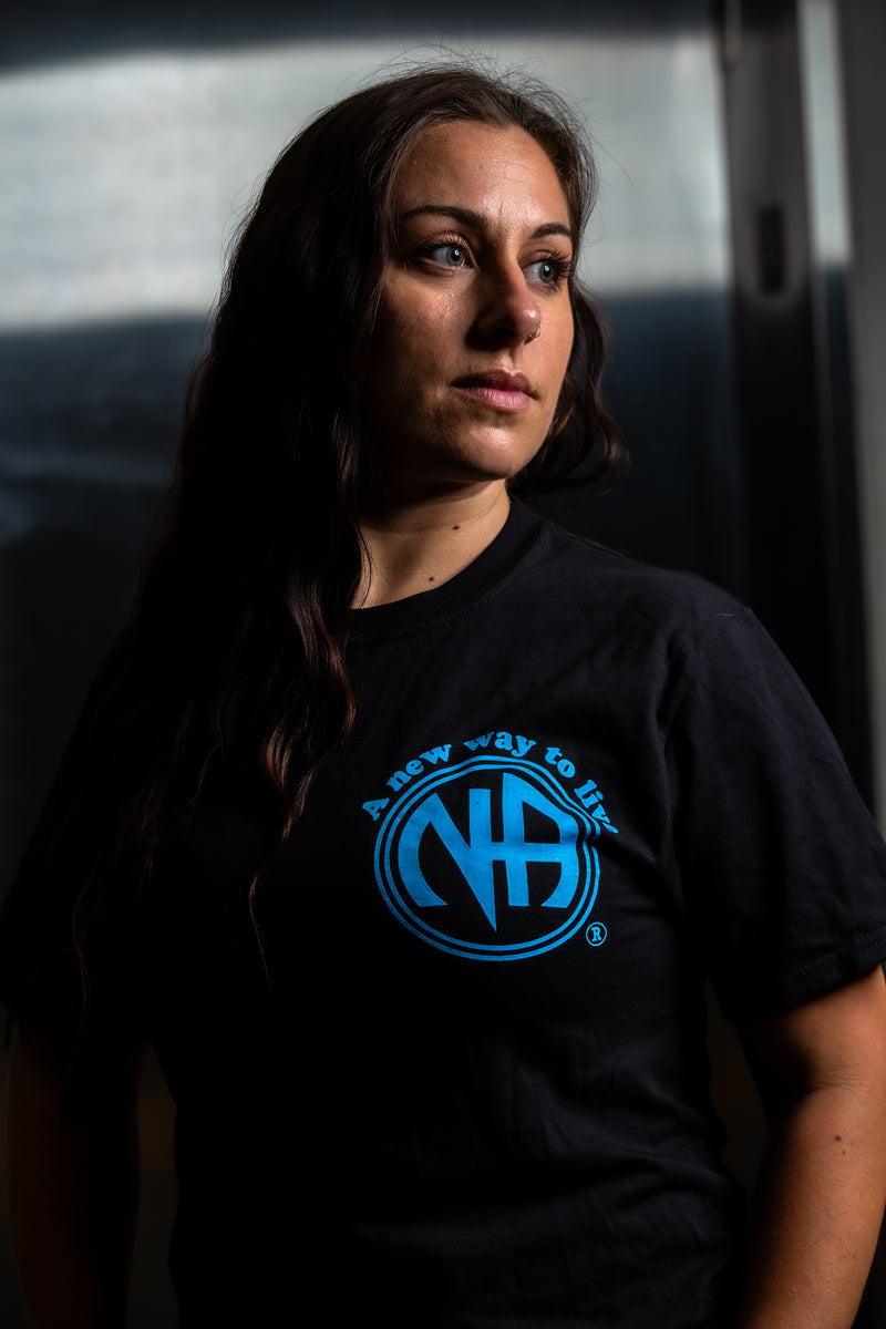 Load image into Gallery viewer, Person wearing a black t-shirt with a blue logo against a dark background
