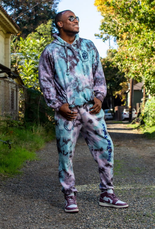 Man wearing a tie-dye hoodie and pants standing on a path with trees in the background