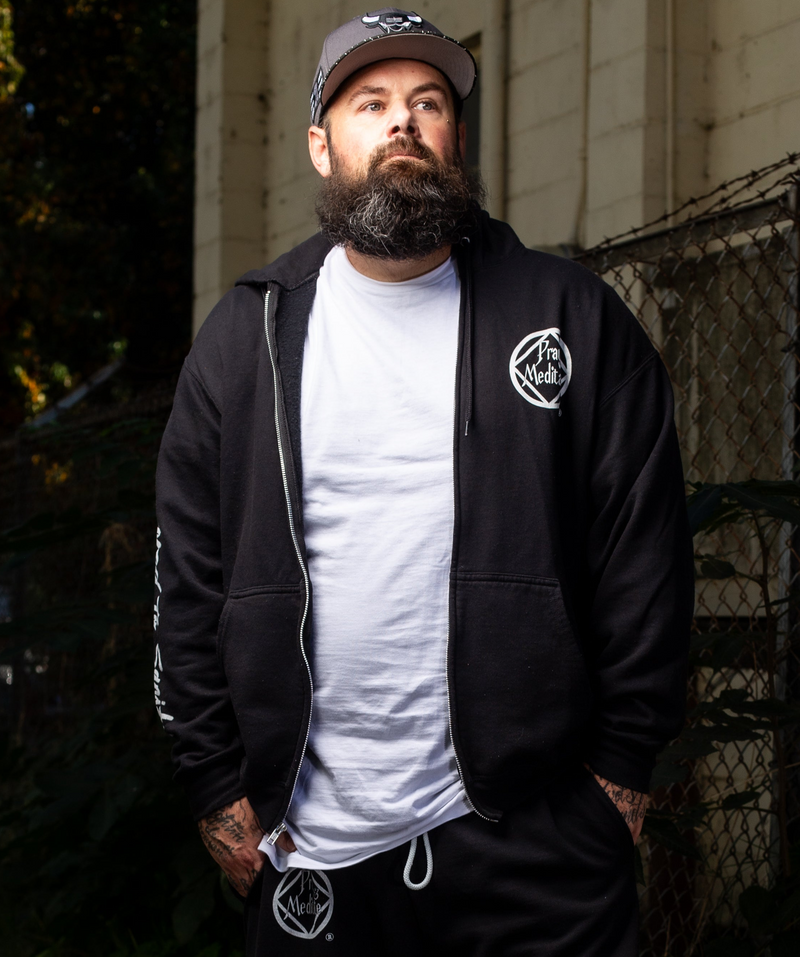 Load image into Gallery viewer, Man wearing a black jacket with a logo, white t-shirt, and dark pants outdoors.
