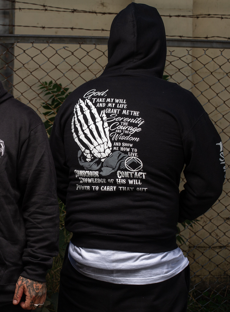 Load image into Gallery viewer, Person wearing a black hoodie with a white graphic and text on the back, standing in front of a chain-link fence.
