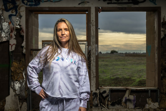 Person wearing a tie-dye outfit standing in an abandoned building with a window showing a field and cloudy sky.