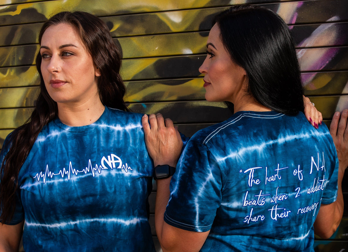 Two women wearing blue tie-dye t-shirts with text against a graffiti-covered wall.