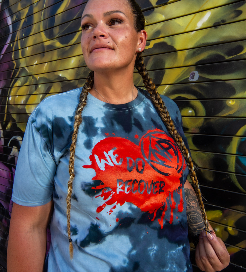 Load image into Gallery viewer, Person wearing a blue tie-dye t-shirt with 'We Do Recover' graphic in front of a colorful mural.
