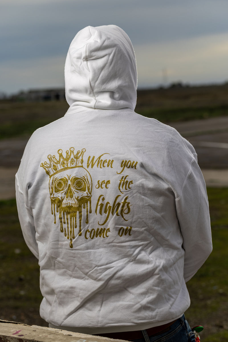 Load image into Gallery viewer, Person wearing a white hoodie with a gold skull design and text on the back, standing outdoors.
