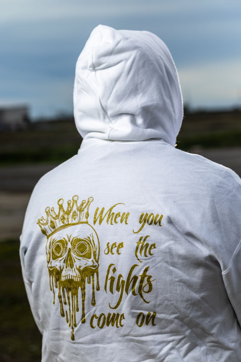Load image into Gallery viewer, White hoodie with gold skull design and text on the back, worn by a person outdoors.
