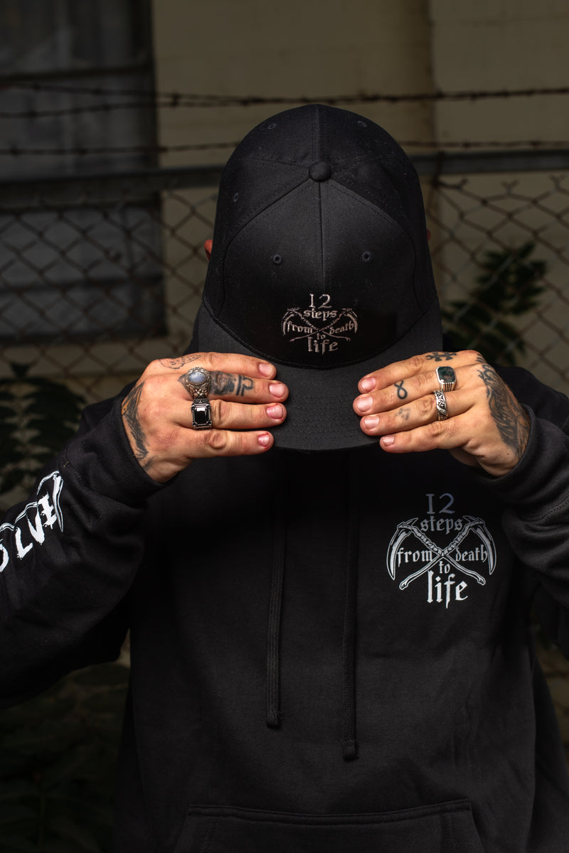 Load image into Gallery viewer, Person wearing a black cap and hoodie with visible branding, holding the cap over their face.
