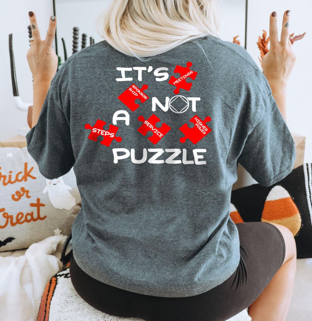 Female model with blonde hair holding up peace signs and wearing a Gildan 64000L in Dark Heather with a full back print that has the words 'It's Not a Puzzle' with puzzle pieces that have text over them with the words 'meetings' 'steps' 'sponsorship' 'service' and 'higher power' and the Narcotics Anonymous Service Symbol