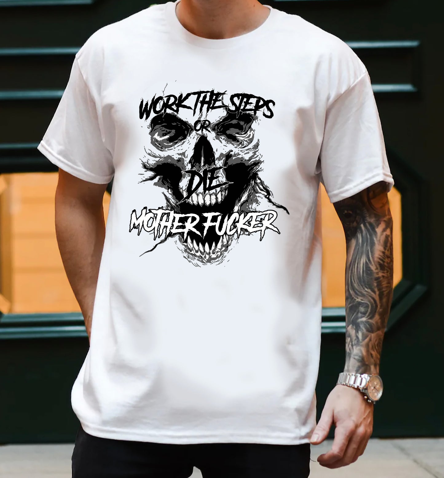 man with tattoo standing in front of a green wall wearing a white tee shirt with a black screaming skeleton skull face with text on top that says "WORK THE STEPS OR DIE MOTHERFUCKER"