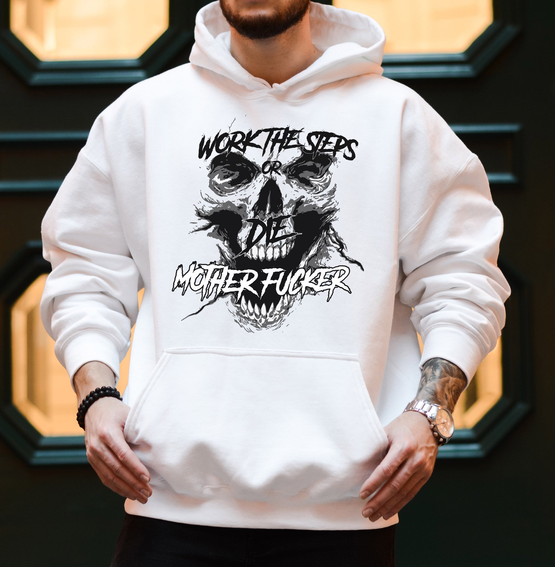 Man with a tattoo standing in front of a green and pale yellow wall wearing a white hooded hoodie/sweatshirt with a black screaming skeleton skull face with text on top that says "WORK THE STEPS OR DIE MOTHERFUCKER" and a front pocket where he is resting his thumbs.